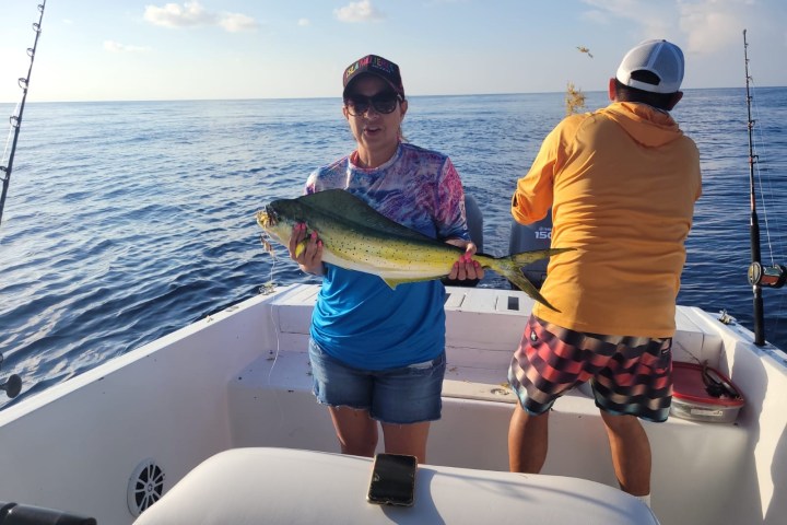 Person holding a large fish on a boat with another person fishing nearby.